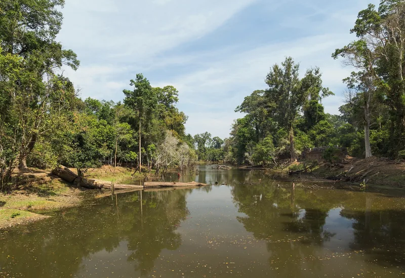 A calm moat lined with dense trees reflects the sky near the ancient temple ruins of Preah Khan in Siem Reap.