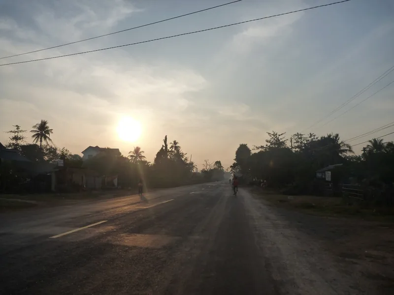 Cyclists ride along a quiet village road near Siem Reap as the sun rises through hazy sky and palm trees.