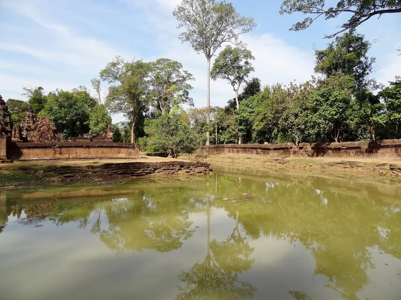 A tranquil moat reflects trees and ancient sandstone walls at Banteay Srei temple near Siem Reap.