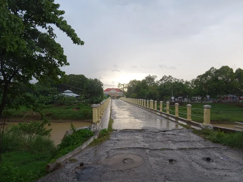 A wet concrete bridge with yellow railings leads toward Battambang's city hall building at dusk.