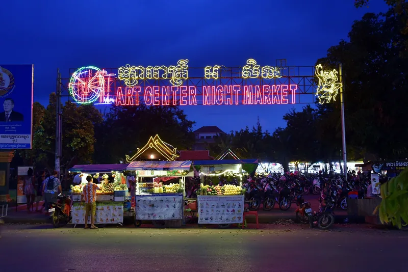 Neon sign lights up the entrance to Art Center Night Market with food stalls at dusk in Siem Reap.