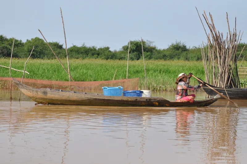 A woman in a wide-brimmed hat paddles a wooden fishing boat with baskets through a brown river near Siem Reap.