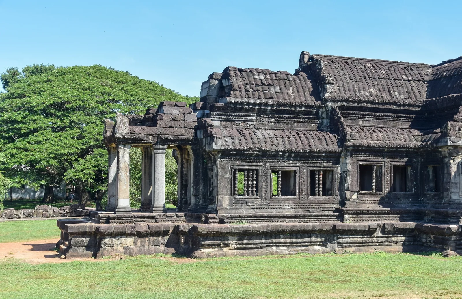 Weathered stone gallery with columns and carved windows at Angkor Wat in Siem Reap, under a clear blue sky.
