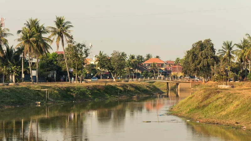 Palm trees line the calm Siem Reap River with a small bridge and town buildings in the background.