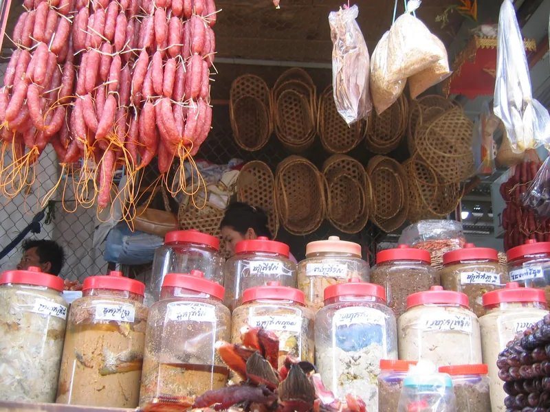 A market stall in Siem Reap displays hanging sausages, woven baskets, and jars of preserved foods with labels.