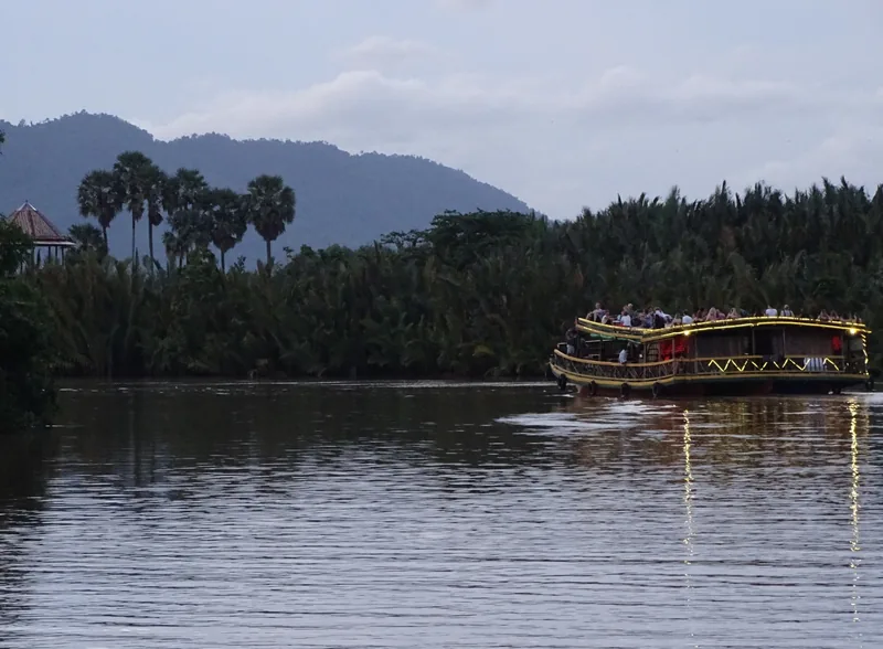 A double-deck tourist boat carries passengers along the Kampot River with palm trees and hills in the background.