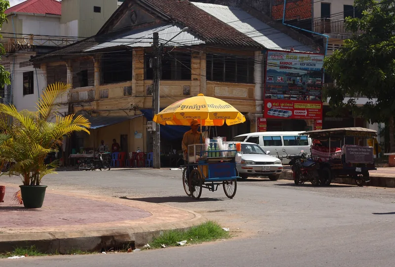 A street vendor with a cart and yellow umbrella stands on a corner in Kampot, Cambodia.