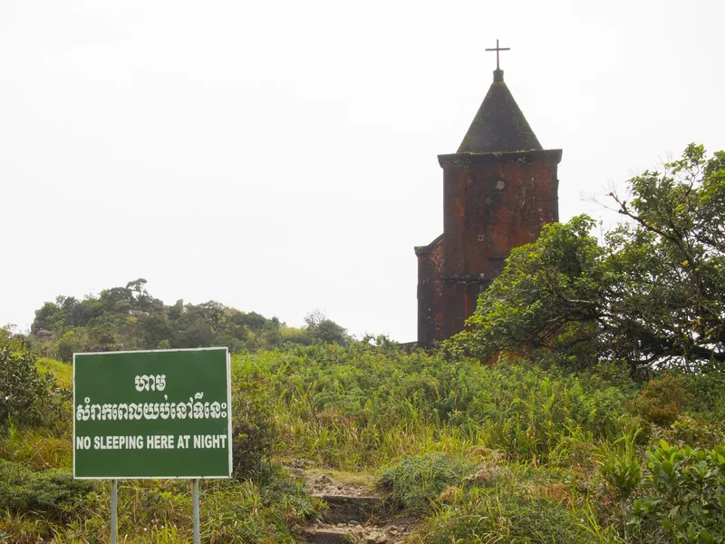 An abandoned church tower stands amid overgrown vegetation on Bokor Hill Station, with a warning sign in the foreground.