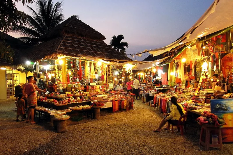 Vendors and shoppers browse a lantern-lit night market with thatched-roof stalls in Siem Reap at dusk.