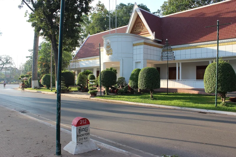 A milestone marking 312 km to Phnom Penh stands before a white building with a red pitched roof in Siem Reap.