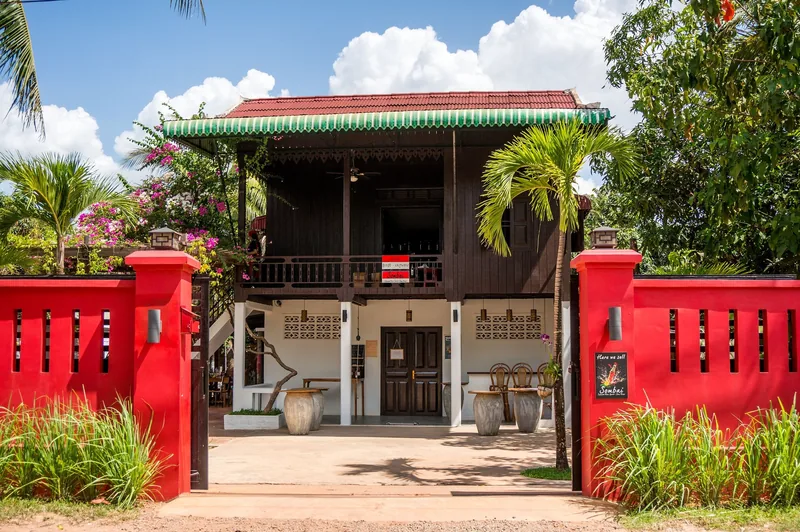 A traditional wooden house with red walls and tropical plants serves as a workshop in Siem Reap, Cambodia.