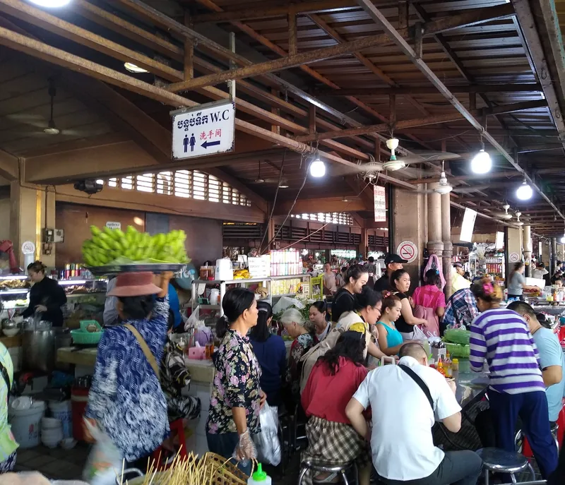 Crowded food stalls under the roof of Phnom Penh's central market with vendors and diners.