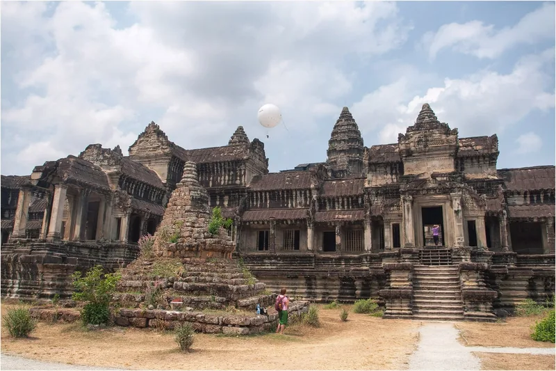 Weathered towers and galleries of Angkor Wat rise above a small stone stupa under a cloudy sky in Siem Reap.