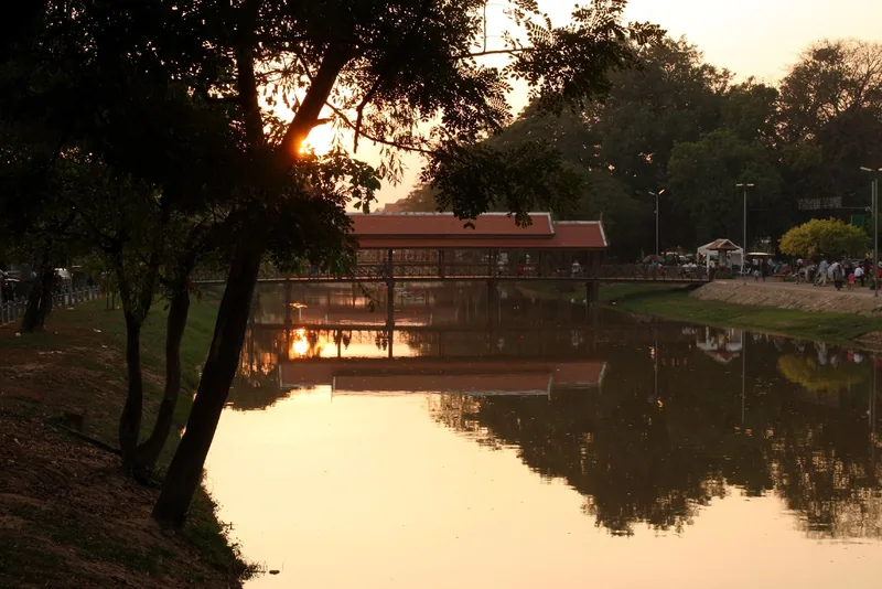 Sunset silhouettes trees beside a covered wooden bridge reflecting in the river at Siem Reap.